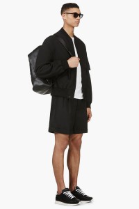 Мужчина в чёрном бомбере Y-3, широких свободных шортах и чёрных кроссовках на белой подошве Kris van Assche, с кожаным рюкзаком 3.1 Phillip Lim, солнечных очках Thom Browne